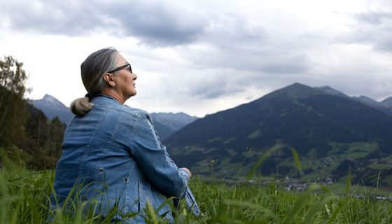 Ältere Frau, sitzt auf einer Wiese und schaut auf die Berge.