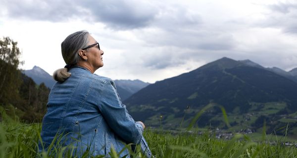 Ältere Frau, sitzt auf einer Wiese und schaut auf die Berge.