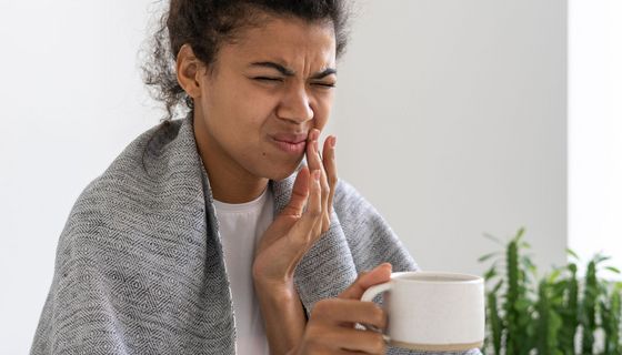 Junge Frau mit Decke und Teetasse verzieht schmerzhaft das Gesicht und fasst sich an die Wange in der Nähe des Mundwinkels.