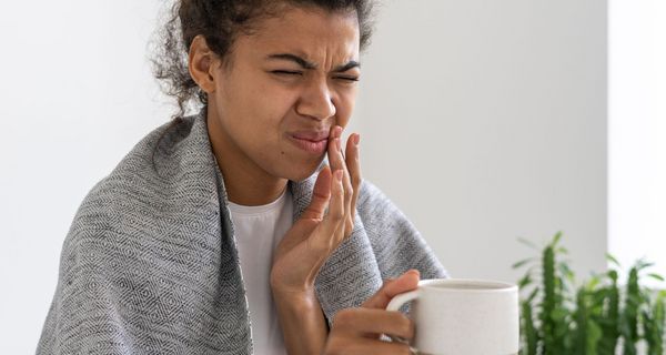 Junge Frau mit Decke und Teetasse verzieht schmerzhaft das Gesicht und fasst sich an die Wange in der Nähe des Mundwinkels.
