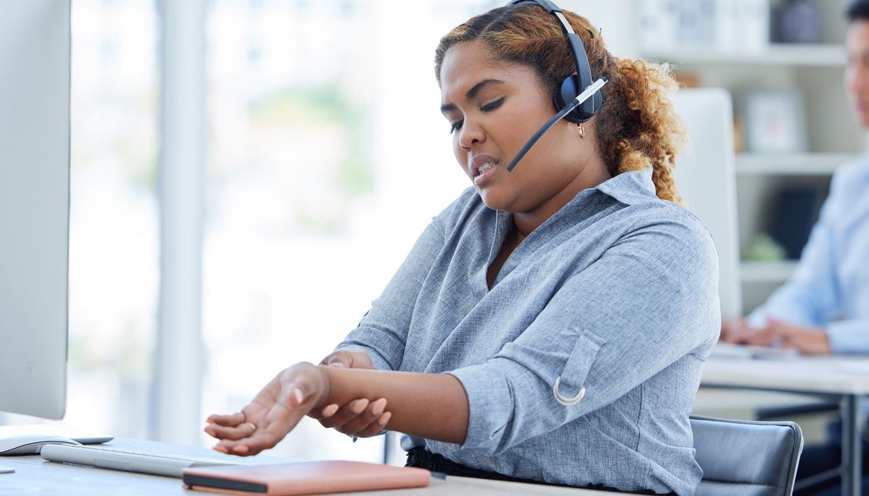 Callcenter? Junge Frau mit Headset sitzt an einem Computer und tastet kritisch das Handgelenk ab.