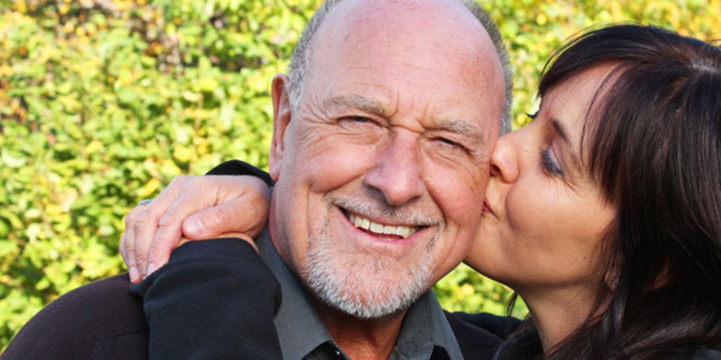 Portrait attraktives Paar Mitte 50; Mann mit grauem Dreitagebart lacht in die Kamera und wird von Frau auf die Wange geküsst