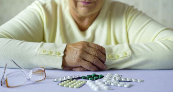 Eine ältere Dame sitzt vor einem Haufen Tabletten in Blistern, ihre Brille daneben. Sie hat die Hände verschränkt.