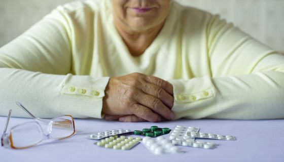 Eine ältere Dame sitzt vor einem Haufen Tabletten in Blistern, ihre Brille daneben. Sie hat die Hände verschränkt.