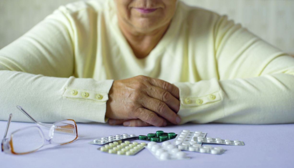 Eine ältere Dame sitzt vor einem Haufen Tabletten in Blistern, ihre Brille daneben. Sie hat die Hände verschränkt.