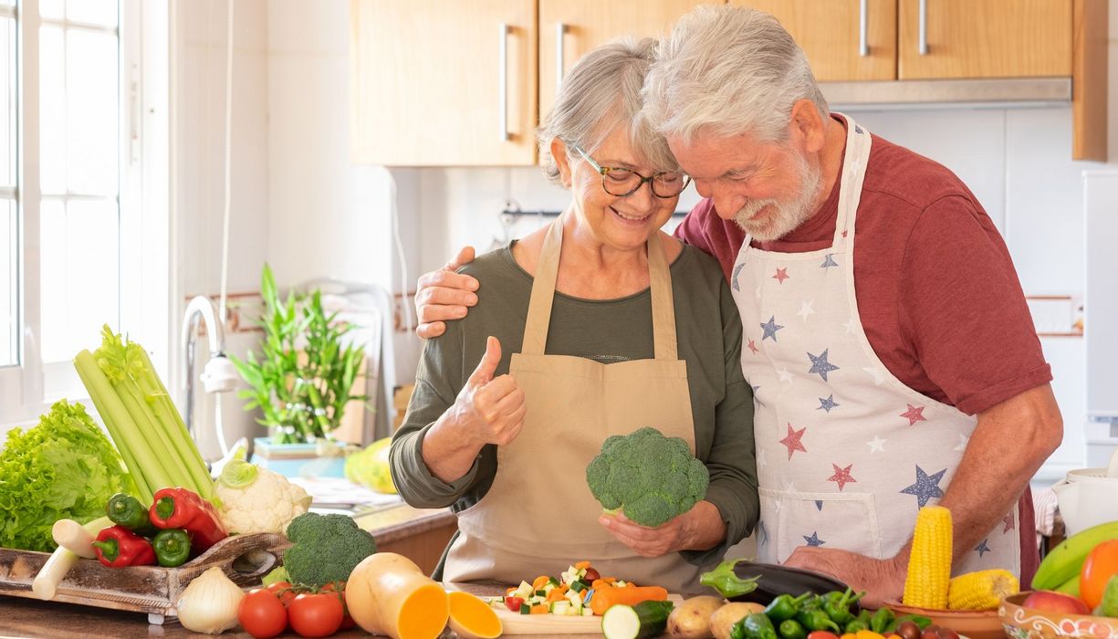 Ein älteres Paar kocht zusammen vegetarisch. Sie schneiden Kürbis, Brokkoli und Mais.