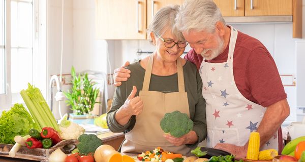 Ein älteres Paar kocht zusammen vegetarisch. Sie schneiden Kürbis, Brokkoli und Mais.
