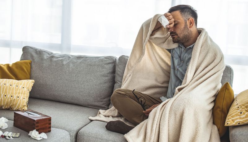 Mann sitzt in eine decke eingewickelt auf dem Sofa, er wischt sich den Schweiß von der Stirn. Um ihn herum liegen benutzte Taschentücher.