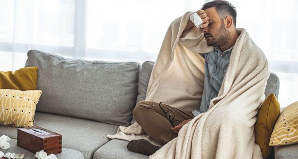 Mann sitzt in eine decke eingewickelt auf dem Sofa, er wischt sich den Schweiß von der Stirn. Um ihn herum liegen benutzte Taschentücher. 