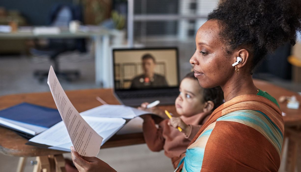 Eine junge Mutter hat ihr Kind auf dem Arm. Sie hält ein Blatt in der Hand und hat Köpfhörer auf. Gleichzeitig läuft im Hintergrund ein Videoanruf.