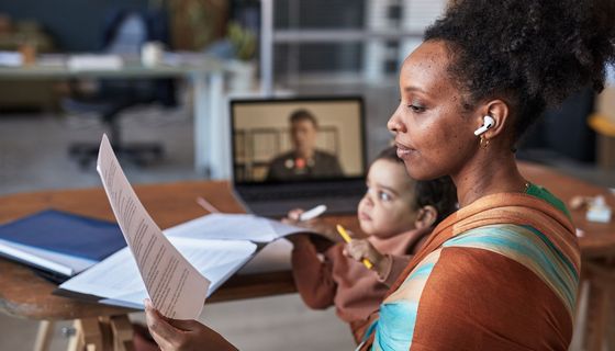 Eine junge Mutter hat ihr Kind auf dem Arm. Sie hält ein Blatt in der Hand und hat Köpfhörer auf. Gleichzeitig läuft im Hintergrund ein Videoanruf.