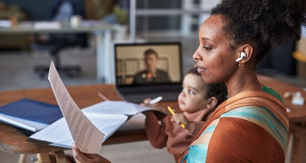 Eine junge Mutter hat ihr Kind auf dem Arm. Sie hält ein Blatt in der Hand und hat Köpfhörer auf. Gleichzeitig läuft im Hintergrund ein Videoanruf.