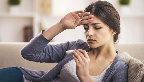 Frau mit Fierber sitzt auf dem Sofa und schaut auf Fieberthermometer