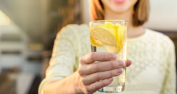 Frau, hält ein Glas mit Wasser und Zitronen in die Kamera.