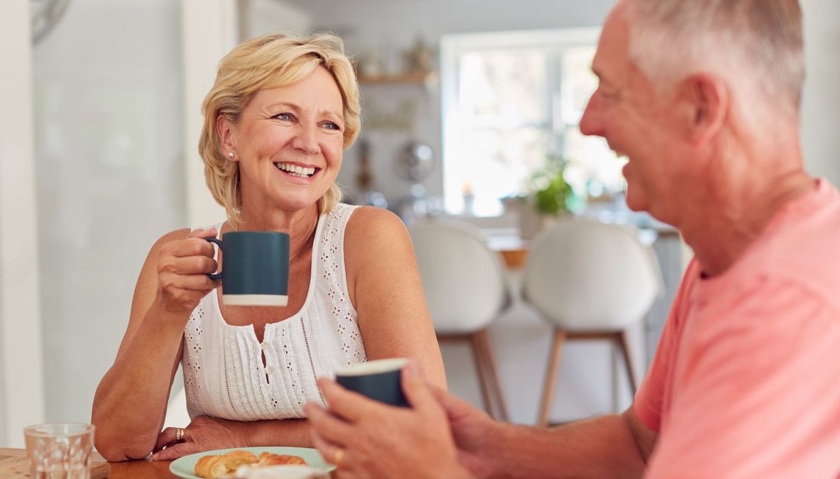 Ein Renter-Paar frühstückt zusammen mit Kaffeetassen, Croissants und Orangensaft. Beide wirken glücklich.