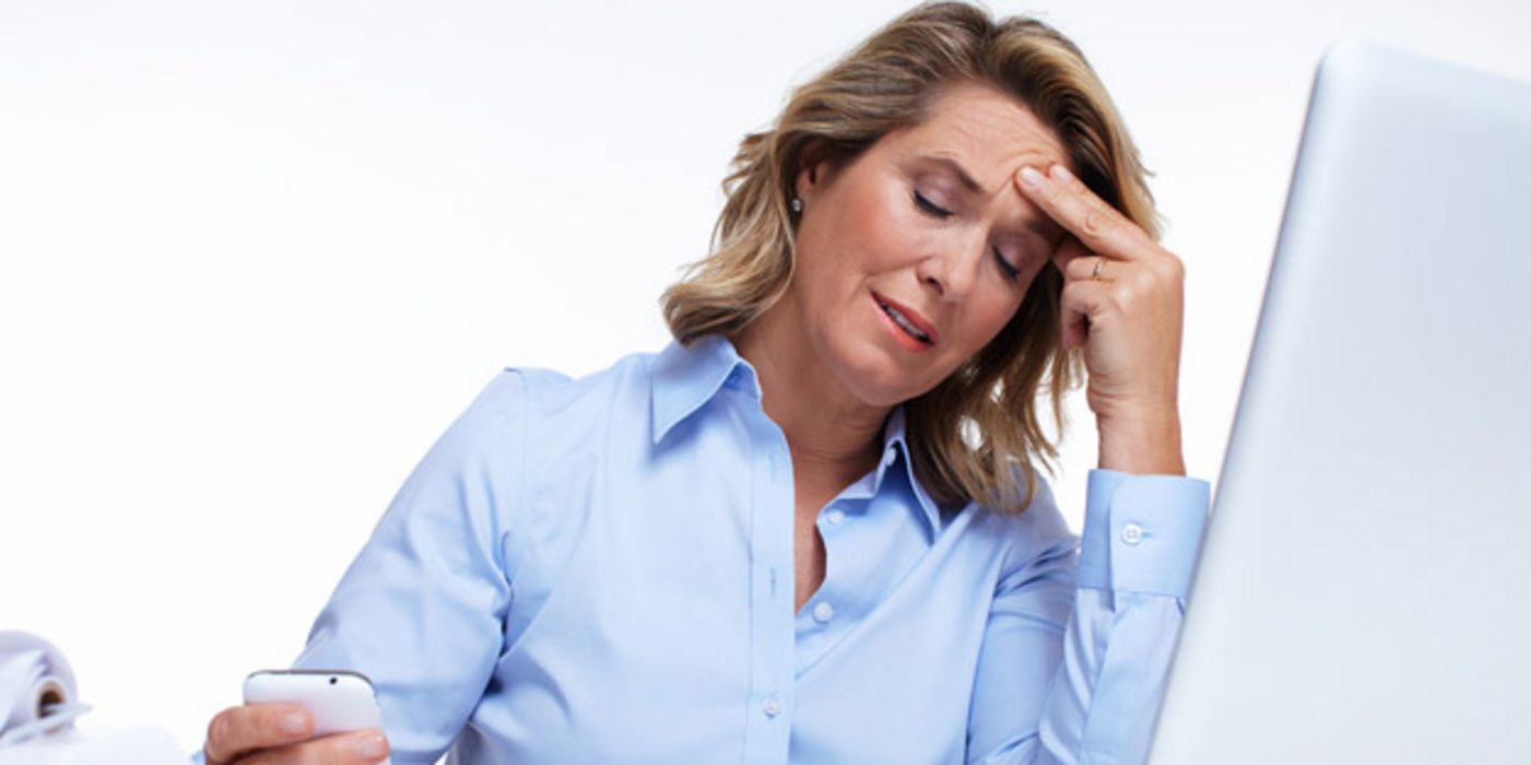 Mittelalte Frau in hellblauer Bluse sitzt vor PC und und fasst sich gestresst an die Stirn, während sie auf ein Smartphone schaut