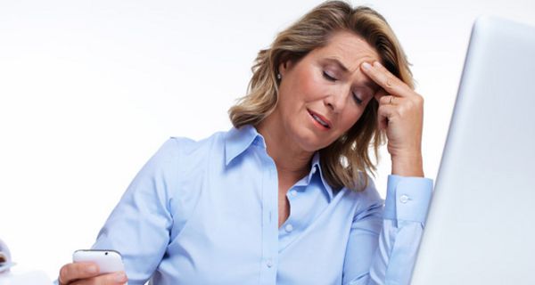 Mittelalte Frau in hellblauer Bluse sitzt vor PC und und fasst sich gestresst an die Stirn, während sie auf ein Smartphone schaut