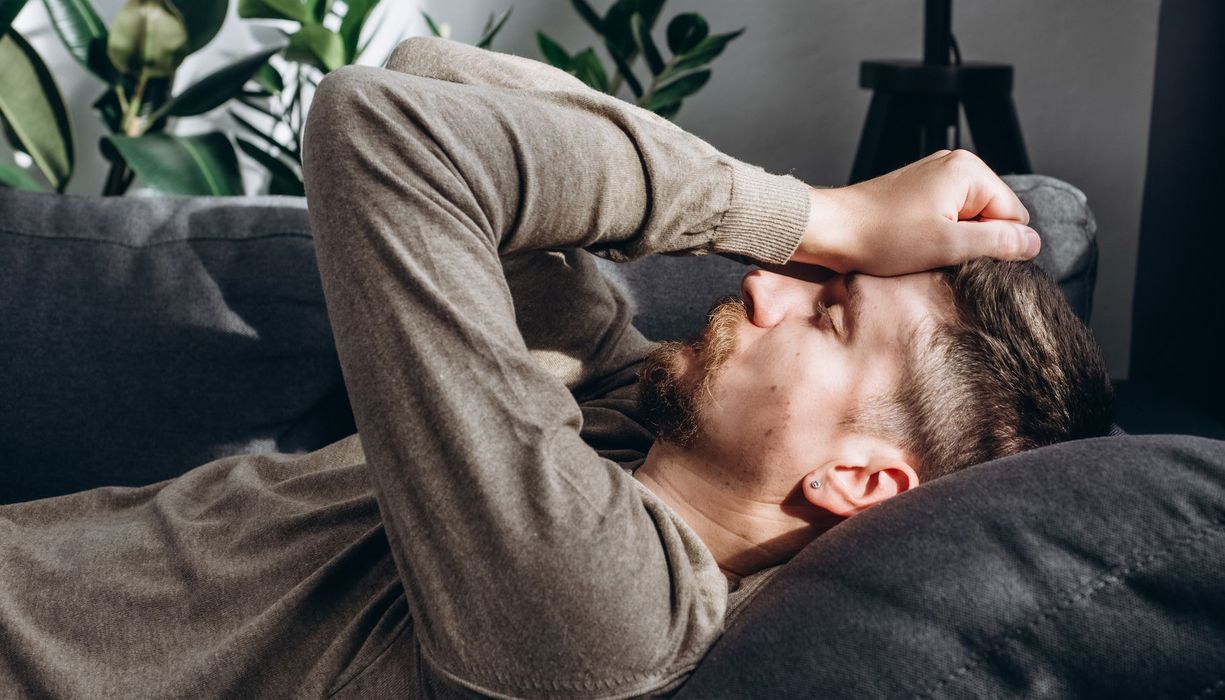 Junger Mann liegt mit geschlossenen Augen auf der Couch, die Daumenballen auf die Stirn gepresst. 