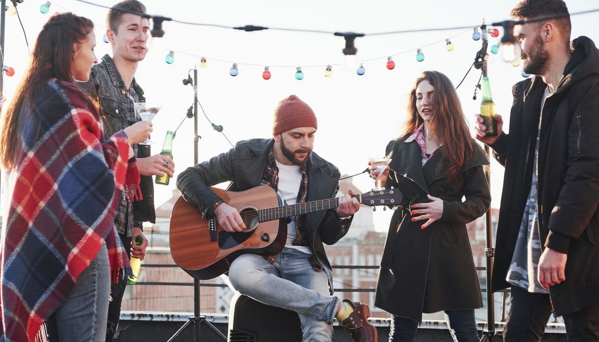 Ein Mann sitzt auf einer Dachterasse und spielt Gitarre. Er ist von zwei Männern und zwei Frauen umgeben, die jeweils ein Getränk in der Hand halten.