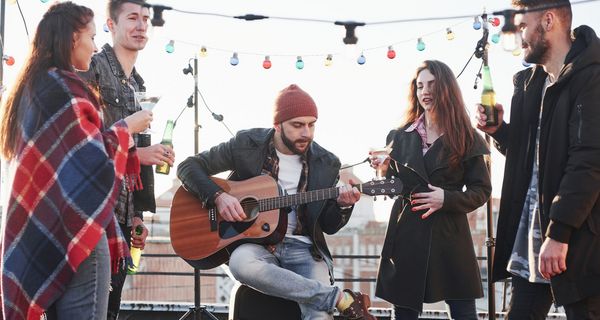 Ein Mann sitzt auf einer Dachterasse und spielt Gitarre. Er ist von zwei Männern und zwei Frauen umgeben, die jeweils ein Getränk in der Hand halten.