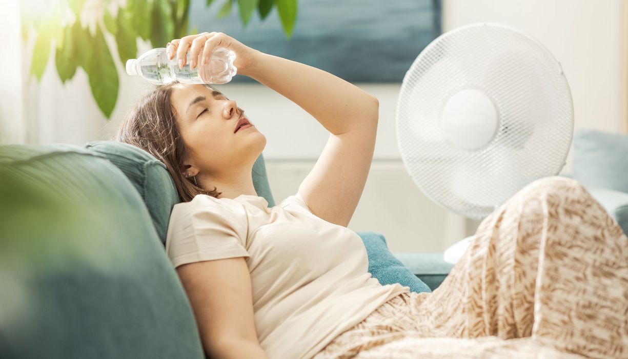 Frau auf dem Sofa hält sich eine kalte Wasserflasche an die Stirn.