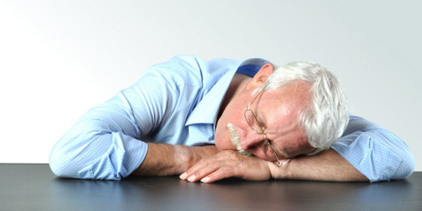 Älterer Mann im hellblauen Hemd schläft am Tisch mit dem Kopf auf den Armen.