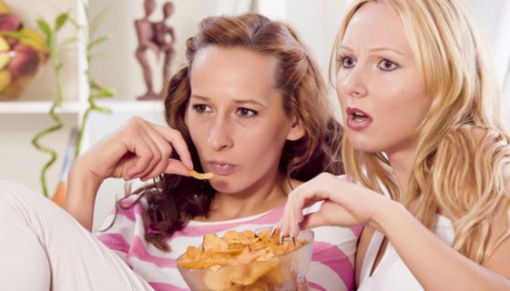 Zwei junge Frauen sitzen auf einer Couch, sehen fern und essen Chips