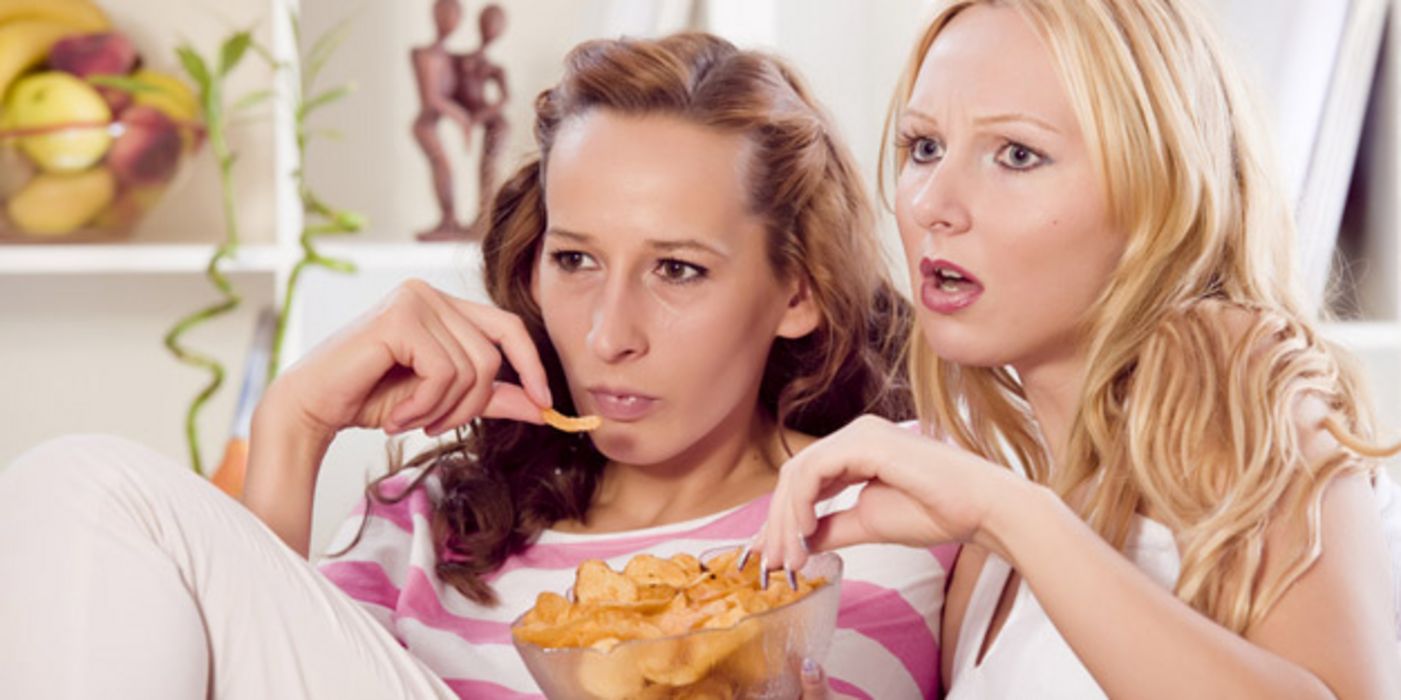 Zwei junge Frauen sitzen auf einer Couch, sehen fern und essen Chips