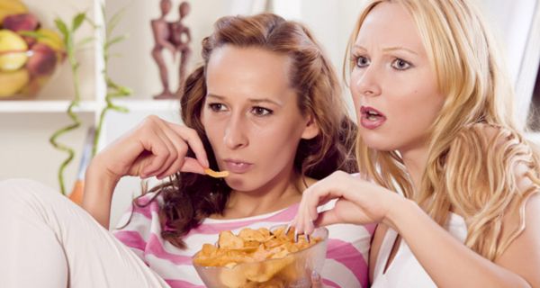 Zwei junge Frauen sitzen auf einer Couch, sehen fern und essen Chips