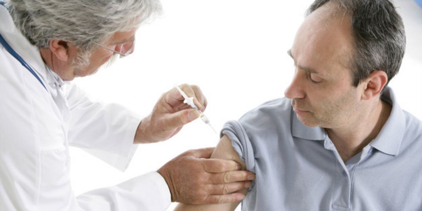 Mann in hellblauem Poloshirt, hochgeschobener Ärmel, schütteres Haar, ca. Mitte 40, bekommt Grippe-Impfung von älterem Arzt