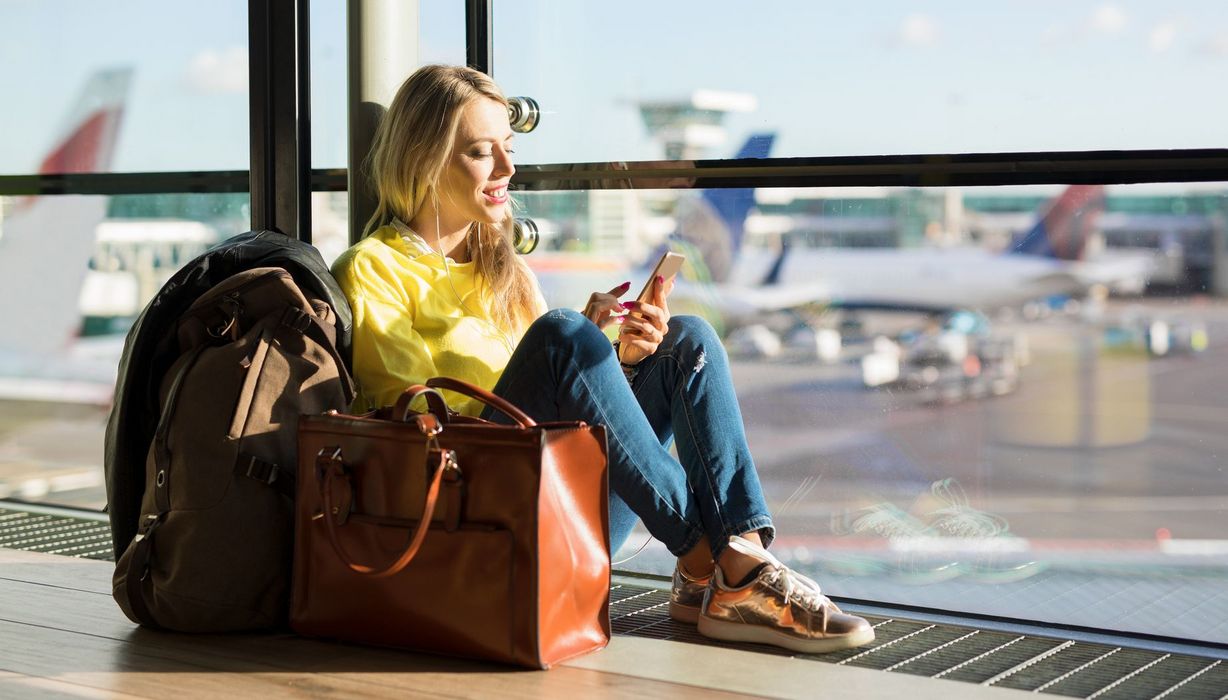 Junge Frau sitzt in der Abflughalle am Fenster. Im Hintergrund sind Flugzeuge zu erkennen.