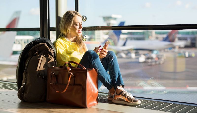 Junge Frau sitzt in der Abflughalle am Fenster. Im Hintergrund sind Flugzeuge zu erkennen.