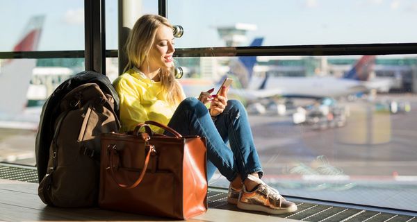 Junge Frau sitzt in der Abflughalle am Fenster. Im Hintergrund sind Flugzeuge zu erkennen.