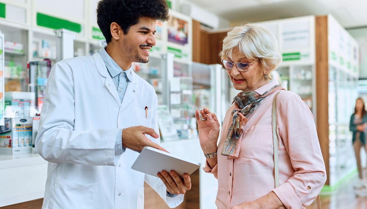 Junger Apotheker erklärt einer älteren Kundin etwas auf einem Tablet.