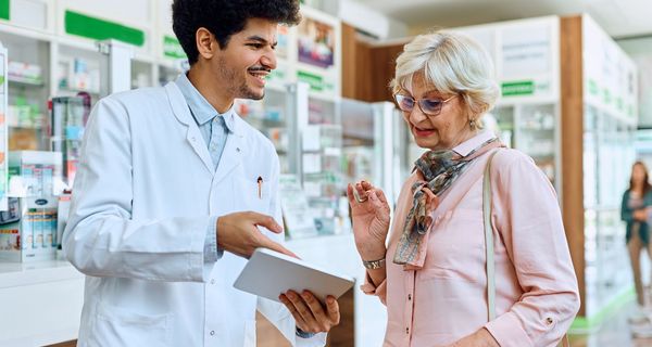 Junger Apotheker erklärt einer älteren Kundin etwas auf einem Tablet.