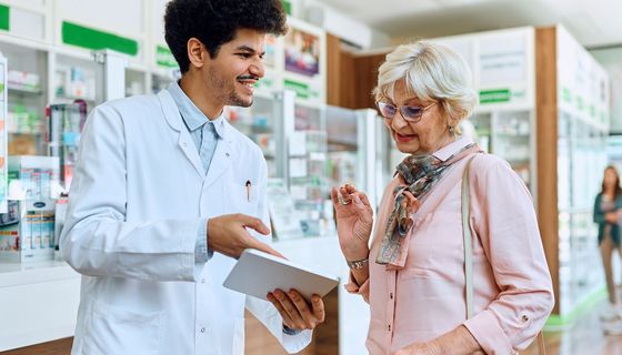 Junger Apotheker erklärt einer älteren Kundin etwas auf einem Tablet.