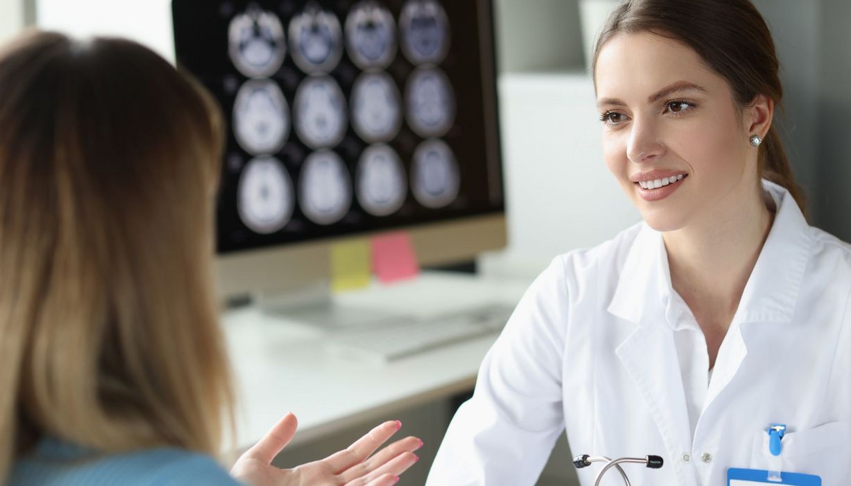 Eine Neurologin berät eine Patientin. Im Hintergrund sind auf einem Bildschirm Hirnscans zu sehen.