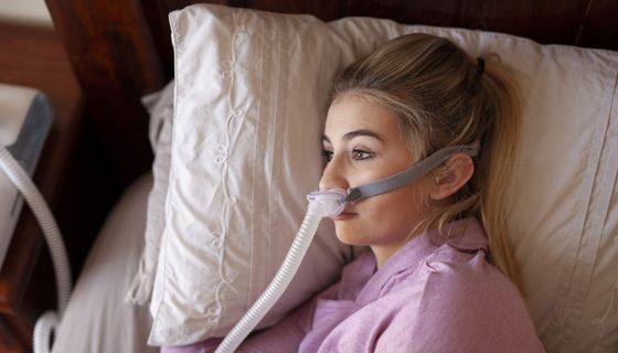 Frau mit Atemmaske liegt im Bett, ihre Augen sind offen.