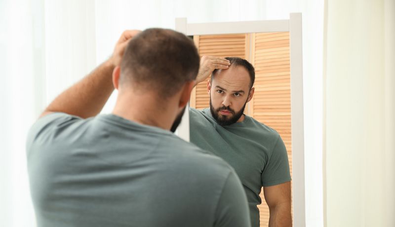 Ein mittelalter MAnn schaut in den Spiegel und betrachtet seinen Haaransatz.