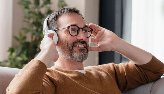 Mann sitzt zufrieden lächelnd auf dem Sofa und fasst sich an seine Kopfhörer.