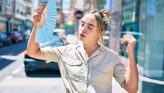 Junge Frau wedelt sich mit einem Fächer Luft zu, hinter ihr ist eine gewöhnliche Straße in der Innenstadt mit Autos und Läden unscharf zu sehen.