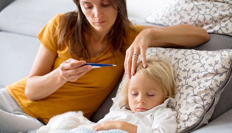 Eine Mutter sitzt auf dem Bett mit ihrem kranken Kind und schaut auf das Fieberthermometer.