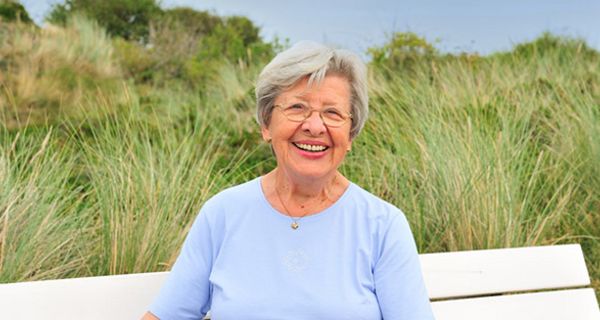 Ältere weißhaarige Dame sitzt auf weißer Bank, im Hintergrund Dünenlandschaft, und lacht in die Kamera