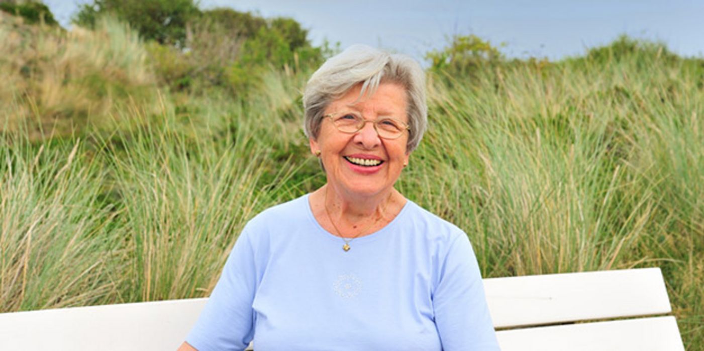 Ältere weißhaarige Dame sitzt auf weißer Bank, im Hintergrund Dünenlandschaft, und lacht in die Kamera