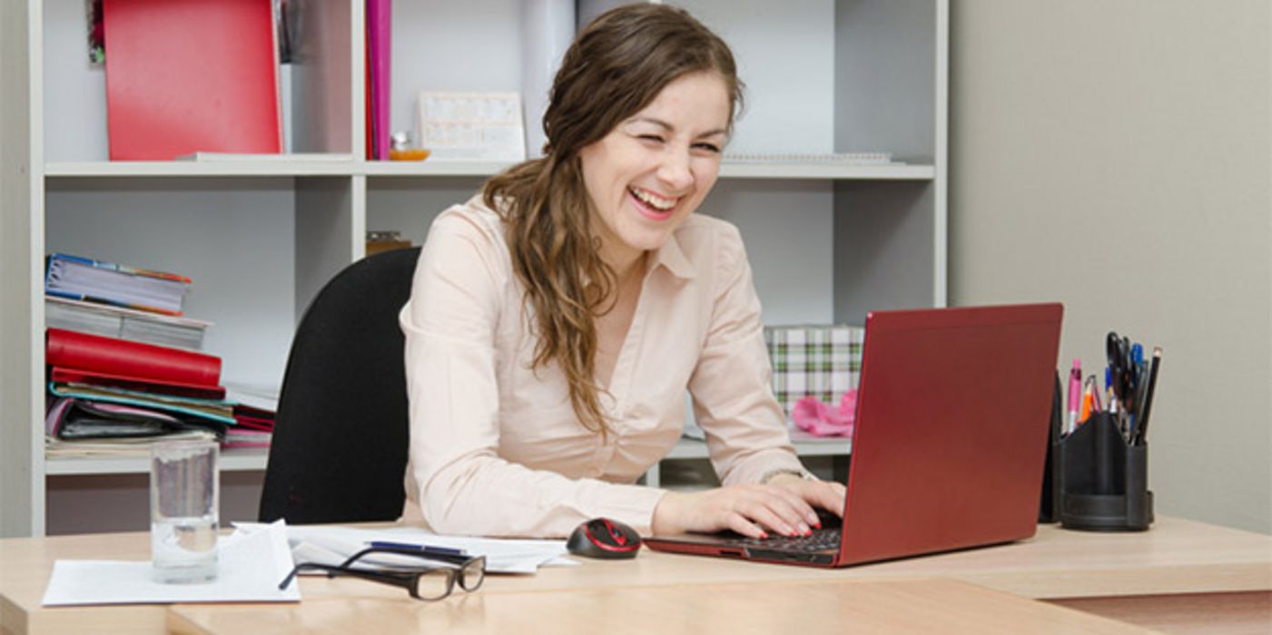 Junge Frau am Schreibtisch schaut herzlich lachend auf ihren Laptop-Bildschirm