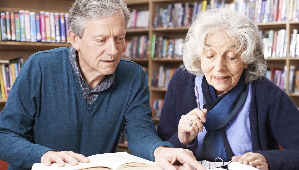 Ein Seniorenpaar liest etwas in Büchern nach. Im Hintergrund sind die Regale einer öffentlichen Bibliothek zu erahnen.