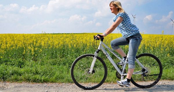 Junge Frau fährt mit dem Fahrrad einen Feldweg entlang; im Hintergrund ein Rapsfeld und Windräder