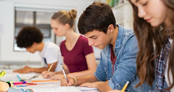 Vier Jugendliche – zwei Mädchen und zwei Jungen – machen in einem Klassenraum Aufgaben aus einem Schulbuch.