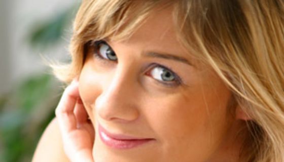 Eine Frau, die Augen dezent geschminkt, Kopf auf die Handfläche gestützt, schaut lächelnd in die Kamera.