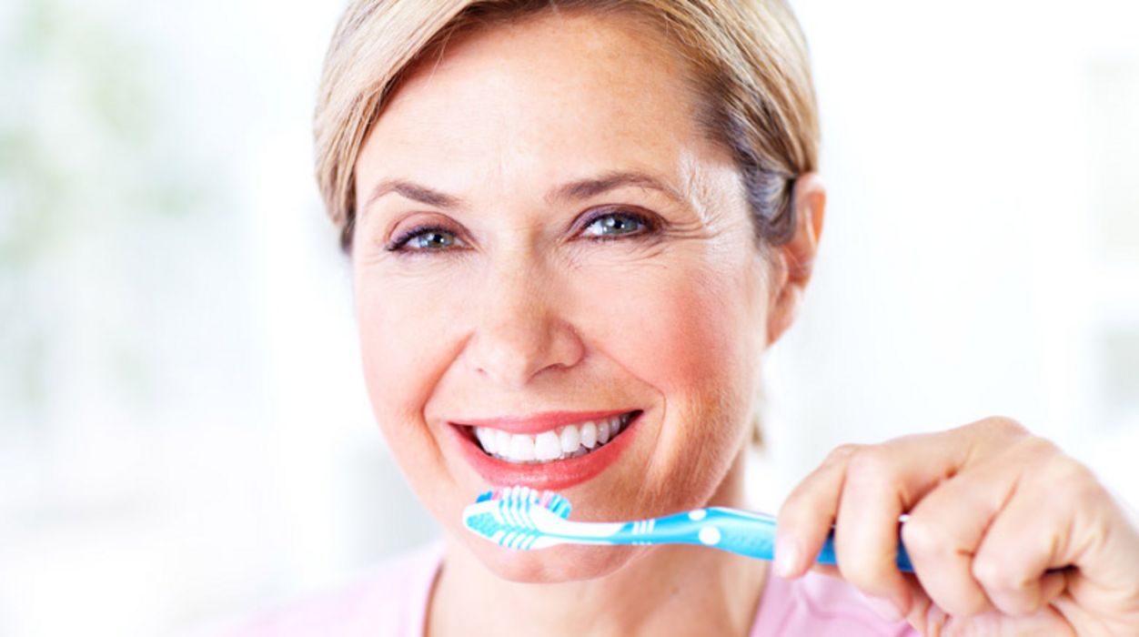 Nahaufnahme: Gesicht einer Frau, Ansatz rosa Pulli, ca. Anfang 50, blond, strahlend lächelnd, schöne Zähne, Zahnbürste in der linken Hand